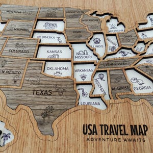 May include: A wooden USA travel map featuring laser-cut state shapes with wood grain textures and white backing. Each state is labelled with its name and a small engraved icon representing local landmarks or culture. The text USA TRAVEL MAP ADVENTURE AWAITS is engraved in the bottom right corner.