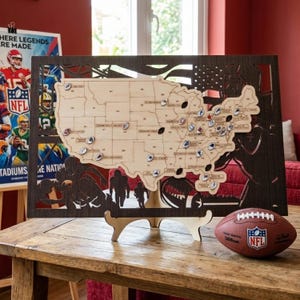 Könnte beinhalten: Eine Holzkarte der Vereinigten Staaten mit Teamlogos, einem dunkelbraunen Rahmen und einem Football. Die Karte wird auf einem Holzständer ausgestellt. Ein NFL-Football liegt auf dem Tisch. Die Karte ist etwa 61 cm breit.