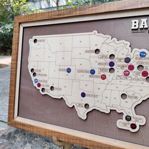 May include: Wooden framed map of the United States with state outlines. The map features baseball stadium markers in various colors. The frame is a dark brown color.
