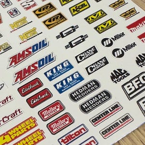 Puede incluir: Una hoja de pegatinas de marcas de carreras y rendimiento automotriz. Las pegatinas presentan varios logotipos en rojo, blanco, negro y azul. Las marcas incluyen AMSOIL, Edelbrock y Hedman Hedders.