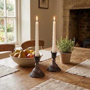 May include: Two lit white spiral candles in ornate bronze candle holders on a wooden table. A bowl of fruit and a potted plant are also on the table. The scene is set in a well-lit room with a window.