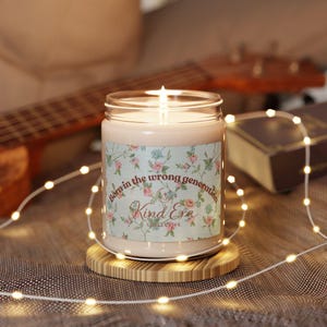 May include: A lit candle in a glass jar with a floral label that reads "Born in the wrong generation" and "Kind Era Collective." The candle sits on a small wooden coaster, surrounded by fairy lights.