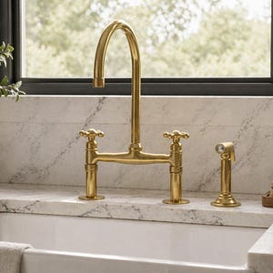 Puede incluir: Un grifo de cocina dorado con un arco alto y manijas en forma de cruz. El grifo está montado en una encimera blanca sobre un fregadero blanco. También se ve un rociador lateral a juego. El fondo incluye una ventana y un salpicadero de mármol.