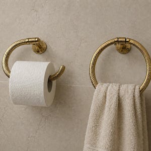 May include: Two gold-colored bathroom fixtures are mounted on a light-colored wall. One is a toilet paper holder with a roll of white paper. The other is a towel ring with a folded white towel. Both fixtures have a textured design.