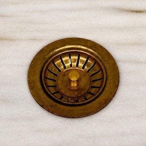 Op de afbeelding: Een close-up van een messing afvoer in een witte marmeren wastafel. De afvoer heeft een cirkelvormig ontwerp met een centrale schroef en een reeks radiale sleuven. Het marmer heeft subtiele grijze en bruine adering.