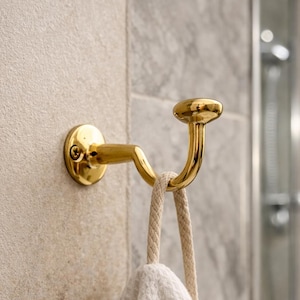 May include: A gold-colored metal towel hook mounted on a beige tiled wall. A white waffle-weave towel hangs from the hook. The hook has a curved design and a round top. The background includes a shower fixture.