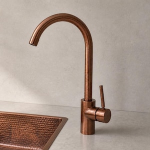 May include: A copper kitchen sink and faucet set. The sink is square with a hammered texture. The faucet has a curved spout and a single lever handle. The set is made of copper and has a warm, reddish-brown color.