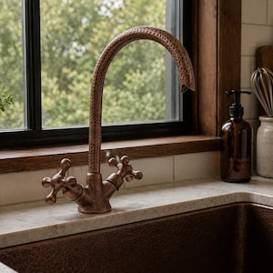 May include: A copper kitchen tap with cross-shaped handles and a curved spout. The tap is mounted on a white countertop above a copper sink. A window with a view of trees is in the background.