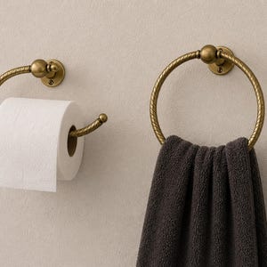 K&ouml;nnte beinhalten: Ein Badezimmer-Set mit einem Toilettenpapierhalter und einem Handtuchring. Beide sind aus antikfarbenem Messingmetall mit einem dekorativen, seilartigen Design gefertigt. Eine Rolle wei&szlig;es Toilettenpapier befindet sich auf dem Halter, und ein dunkelgraues Handtuch h&auml;ngt am Ring.