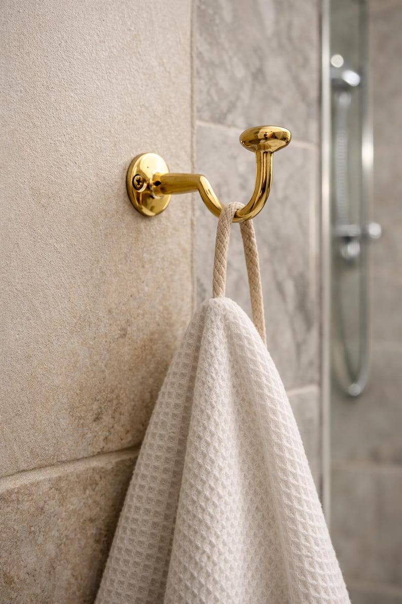 Pu&ograve; includere: Un gancio portasciugamani in metallo color oro montato su una parete piastrellata beige. Un asciugamano bianco a trama waffle &egrave; appeso al gancio. Il gancio ha un design curvo e una parte superiore rotonda. Sullo sfondo, un soffione doccia.