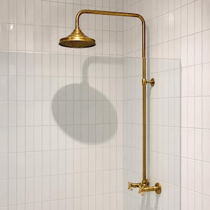 Puede incluir: Un conjunto de ducha de estilo vintage con acabado de latón. El cabezal de la ducha es redondo y el grifo tiene manijas en forma de cruz. El conjunto está montado en una pared de azulejos blancos. La sombra del cabezal de la ducha es visible.