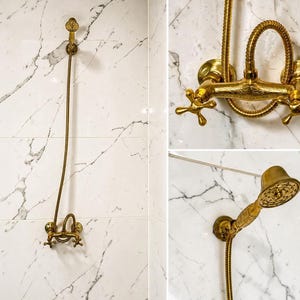 Könnte beinhalten: Ein goldfarbenes Duscharmaturen-Set vor einer weiß marmorierten Wand. Das Set umfasst einen Duschkopf, einen Wasserhahn und eine Handbrause, alle mit aufwendigen Details. Die Armaturen haben eine Vintage- oder Antik-Ästhetik.