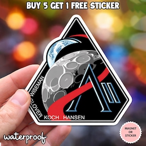 Puede incluir: Una pegatina triangular negra con un diseño estilizado de la luna y la Tierra, con los nombres "WISEMAN", "SLOVER", "KOCH" y "HANSEN". La pegatina también tiene el texto "waterproof" y "MAGNET OR STICKER".