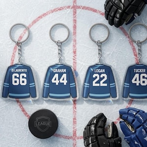 Op de afbeelding: Vier hockeytrui sleutelhangers met spelersnamen en nummers: Di Laurentis 66, Graham 44, Logan 22 en Tucker 46. Elke sleutelhanger is blauw met witte accenten. Een zwarte hockeypuck en handschoenen zijn ook zichtbaar.