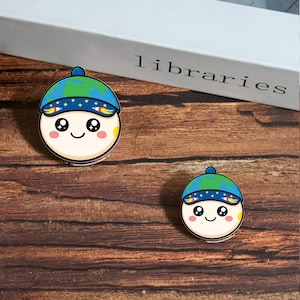 Puede incluir: Dos pegatinas redondas de estilo caricatura con una cara sonriente que lleva un sombrero azul y verde con un diseño de globo terráqueo. Las pegatinas tienen un fondo blanco con mejillas rosas y un acento amarillo. La palabra "libraries" es parcialmente visible.