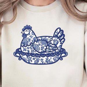 May include: Cream-colored sweatshirt featuring a blue and white decorative hen design. The hen is resting in a nest with floral patterns. The sweatshirt is a casual style, suitable for everyday wear.