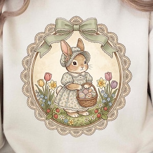 May include: A cream-colored sweatshirt featuring a circular illustration of an Easter bunny holding a basket of eggs. The bunny is wearing a bonnet and dress, surrounded by flowers and a green ribbon.