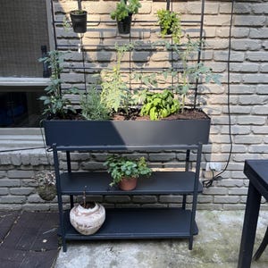May include: A black metal outdoor planter with three shelves and a trellis. The top shelf holds a rectangular planter box with various plants. The trellis supports hanging pots and climbing plants. A decorative pot and a hanging plant are also visible.