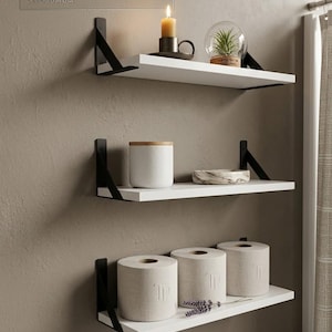 May include: A modern bathroom shelf set with three white shelves supported by black metal brackets. The shelves hold a candle, a small plant in a glass globe, a white jar, a decorative bowl, and rolls of toilet paper. Text reads "ELEVATE YOUR SPACE".