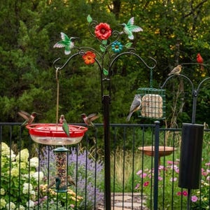 May include: A black metal bird feeder stand with a red hummingbird feeder, a suet feeder, and a bird bath. The stand is decorated with colorful metal flowers and hummingbirds. Several birds are perched on the feeders.