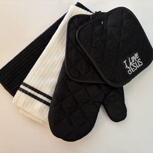 May include: A set of kitchen textiles including a black quilted oven mitt, a black quilted pot holder with the white embroidered text I LOVE JESUS, and two ribbed kitchen towels, one in solid black and one in white with two black stripes.