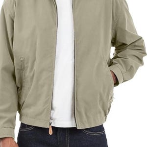 May include: A tan zip-up jacket with a collared neckline and two front pockets. The jacket has a zipper closure and a small leather pull. The person is wearing a white t-shirt and dark blue jeans.