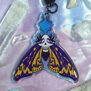 May include: A moth-shaped acrylic keychain featuring a central white skull with purple eyes and black crown-like details. The wings are colored in deep purple and vibrant yellow with a holographic finish, attached to a black metal lobster clasp.