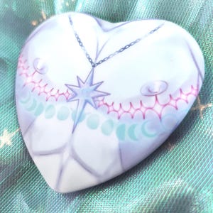 May include: A white heart-shaped ceramic ornament with a silver chain and a star pendant. The ornament is decorated with a pink and green design.