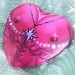 May include: A pink heart-shaped resin ornament with a blue star and chain design. The ornament has a white and blue design on the front.
