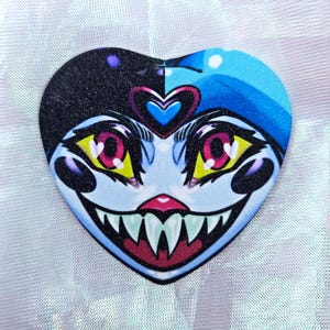 May include: A heart-shaped patch with a blue and black background featuring a cartoon face with yellow eyes, pink eyelids, and a large, toothy grin. The face has a blue and pink heart on its forehead.