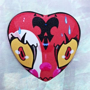 May include: A red heart-shaped pin with a black and blue design. The design features a stylized heart with a black butterfly inside. The heart has a white outline and two yellow eyes with black pupils. The heart is covered in glitter.