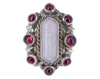 May include: A silver-toned ring with a central, elongated, white opal stone. The opal is surrounded by a twisted rope design and framed by nine round, deep red garnet stones. The ring has an ornate, vintage aesthetic.