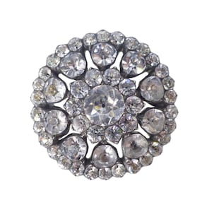 Puede incluir: Un broche redondo con un engaste de plata y un racimo de piedras transparentes y facetadas.