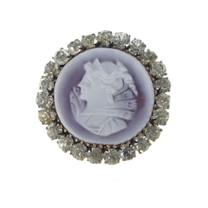 May include: A round cameo brooch with a white cameo of a woman's profile surrounded by a circle of clear rhinestones.