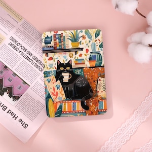 Può includere: Un oggetto rettangolare decorativo con un gatto nero che tiene una tazza, seduto su un divano colorato. Lo sfondo include una libreria con libri e piante. Il testo "YOUNG FLOWER IN A DESERT" è visibile.