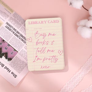 May include: A rectangular library card with the text "Buy me books & Tell me I'm pretty xoxo" in pink script. The card has a lined paper design and the words "LIBRARY CARD" at the top. A newspaper and lace trim are in the background.