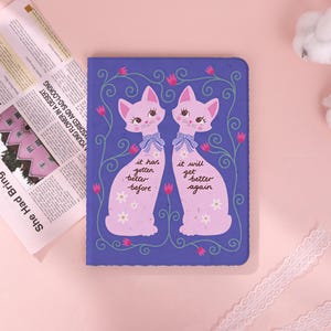 May include: A rectangular purple notebook featuring two light pink cats with blue neck ribbons and white daisy patterns. The text reads, "it has gotten better before" and "it will get better again," surrounded by green vines and pink tulips.