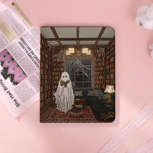 May include: A rectangular notebook cover featuring a printed illustration of a ghost reading a book inside a library with bookshelves, a chandelier, a dark sofa, a patterned rug, and spiderwebs on the walls and ceiling.
