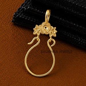 Könnte beinhalten: Ein goldfarbener Metallanhänger mit einem Blumenmotiv an der Oberseite, einer zentralen kreisförmigen Öffnung und einer hakenförmigen Öse. Das Design umfasst kleine blattartige Details um die Blume herum, mit dem Text SHUBHARA JEWELS.