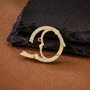 Könnte beinhalten: Ein goldener kreisförmiger Anhänger mit einem aufklappbaren Mechanismus, verziert mit kleinen klaren Steinen am äußeren Rand und einem einzelnen kleinen roten Stein. Das Schmuckstück trägt die Gravur SHUBHRA JEWEL auf der Oberfläche.