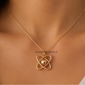 May include: A gold-toned necklace featuring a pendant shaped like an atom with two intersecting elliptical orbits surrounding a central sphere with a faceted, textured surface. The text SHUBHRA JEWELS is visible above the pendant.