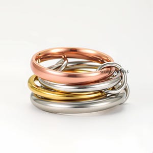 Könnte beinhalten: Ein Stapel von fünf Ringen in verschiedenen Metallausführungen: Roségold, Gold, Silber und ein verbundenes Silberdesign. Die Ringe sind übereinander gestapelt, wodurch ein geschichteter Effekt entsteht. Die Ringe sind auf weißem Hintergrund.