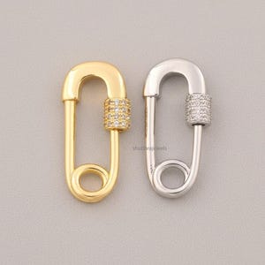 Könnte beinhalten: Zwei dekorative Sicherheitsnadel-Anhänger, einer goldfarben und einer silberfarben, werden vor einem grauen Hintergrund gezeigt. Die goldene Nadel hat einen Zylinder aus kleinen, klaren Steinen. Die silberne Nadel hat einen ähnlichen, mit Steinen besetzten Zylinder.