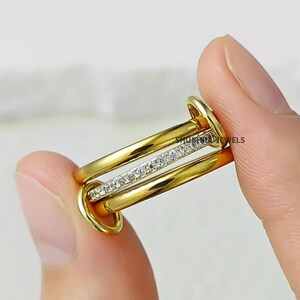 Könnte beinhalten: Ein goldfarbener Ring mit drei ineinandergreifenden Bändern. Ein Band ist mit kleinen, klaren Edelsteinen besetzt. Die Ringe werden von den Fingern einer Person gehalten. Der Text "SHUBHJEWELS" ist sichtbar.