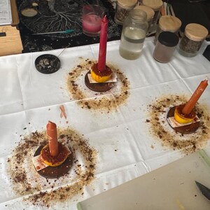 Può includere: Una tovaglia bianca con tre candele, ciascuna sopra una piccola base di legno con un accento giallo e marrone. Le candele sono rosse e arancioni. Intorno a ogni candela c'è un motivo circolare di polvere marrone. Vari barattoli e contenitori sono sullo sfondo.