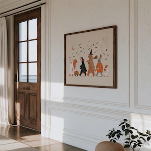 May include: A framed wall art print featuring a whimsical Halloween illustration of a squirrel, badger, bear, rabbit, and mouse dressed in costumes. The animals are walking together under a crescent moon and a swarm of black bats on a beige background.
