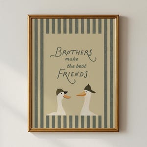 Może przedstawiać: Oprawiony wydruk ścienny przedstawiający dwie białe gęsi w ciemnych czapkach, umieszczone pod napisem "Brothers make the best FRIENDS" ciemną czcionką. Wzór znajduje się na beżowym tle z ciemnoszarymi pionowymi pasami na obrzeżach.