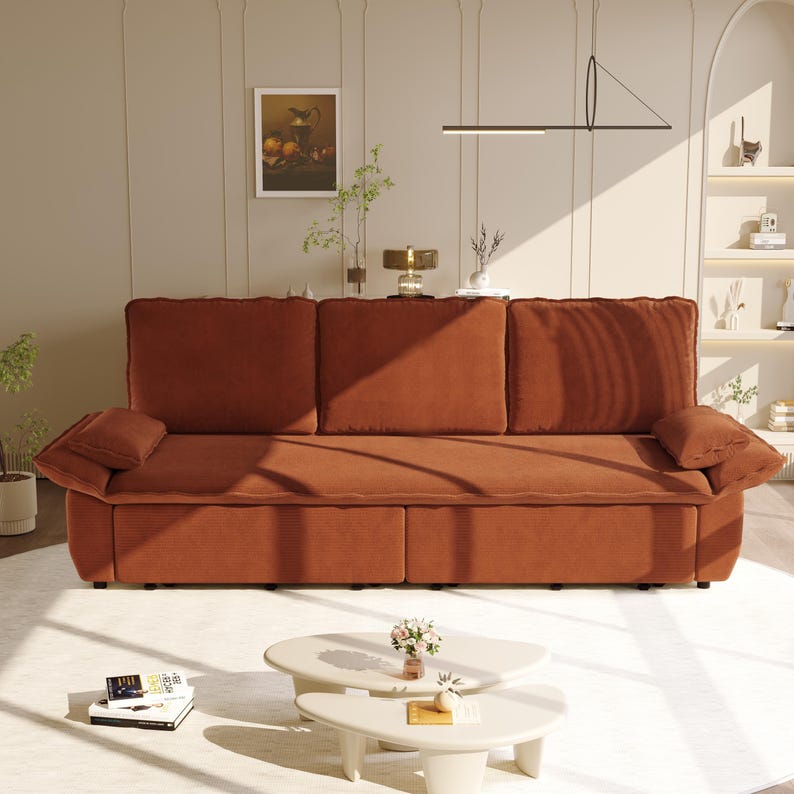 May include: A large, three-cushion sofa in a warm, reddish-brown hue. The sofa features two armrests and several throw pillows. The room is decorated with a painting, a lamp, and two small, white, oval-shaped coffee tables.