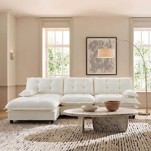 May include: A white sectional sofa with tufted cushions and a chaise lounge. A marble coffee table with a woven basket and a bowl sits on a patterned rug. A floor lamp with a beige shade and a framed abstract painting complete the living room setting.