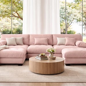 May include: A large, modular sectional sofa upholstered in soft pink fabric, featuring tufted seat cushions, two matching ottomans on the ends, and several throw pillows in cream and pink tones, arranged around a round, light-toned wooden coffee table.